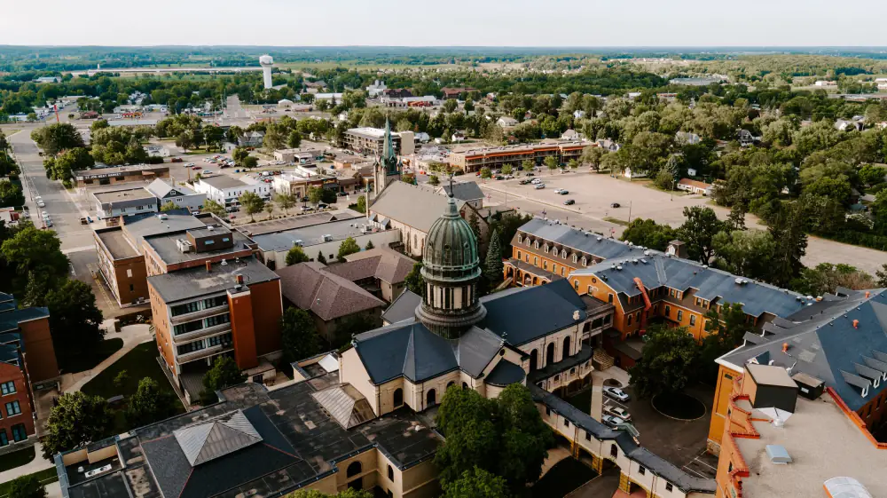 Downtown St. Joseph, Minnesota from the air: the abbey church and its green copper dome at the centre, low brick storefronts and parking lots along the streets around it, and trees running out to the horizon.
